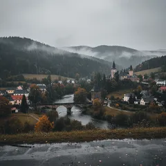 Podzimní krajina české vesnice s kostelem, mostem a mlhavými kopci v pozadí.