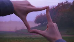 Ruce tvořící rámeček prsty před krajinou při západu slunce.