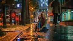 Žena sedí na mokrém chodníku v nočním městě, osvětleném pouličními lampami.
