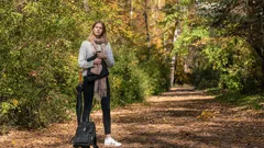 Žena s kufrem stojí na podzimní lesní cestě, oblečená v šále a bundě.