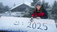 Žena píše 2025 do sněhu na střeše v zimní krajině, úsměvavý obličej, zima, smrky.