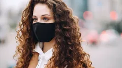 Žena s kudrnatými vlasy a černou rouškou venku ve městě během pandemie.