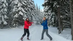 Dva lidé se baví na sněhu v zimním lese, obklopeni zasněženými stromy.