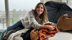 Usměvavá žena leží na posteli s oblečením, v pozadí deštník a taška, domácí prostředí.