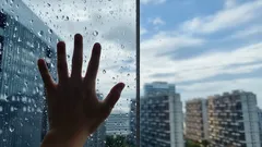 Ruka na okně s dešťovými kapkami, výhled na město a mrakodrapy za sklem